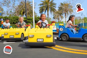 A group of young children ride colorful, LEGO-style cars at an amusement park driving school attraction. The kids are smiling and laughing as they drive yellow and blue toy vehicles along a small road, with palm trees and playful LEGO decorations in the background. The scene is lively and cheerful, with animated motion and stickers of steering wheels and LEGO heads enhancing the fun atmosphere.